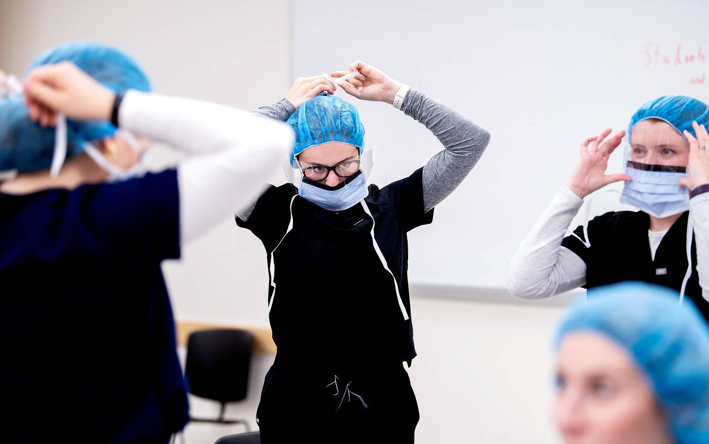 Several individuals in a clinical setting put on blue surgical caps and face masks while wearing scrubs.